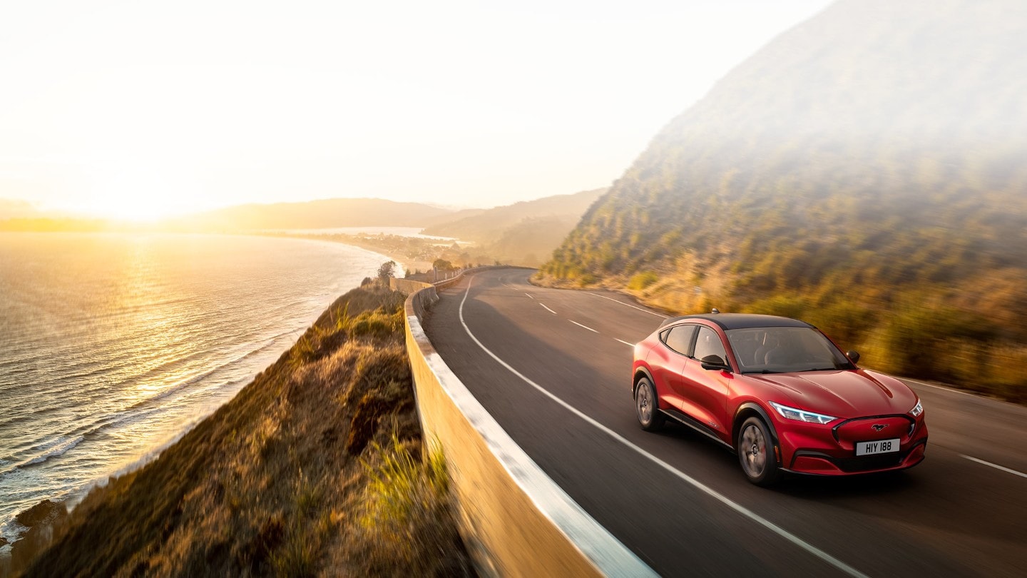 Ford vollelektrische Fahrzeuge. Ford Mustang Mach-E in Rot. Seitenansicht, fahrend auf Straße an Meer mit baumbewachsenen Ufern
