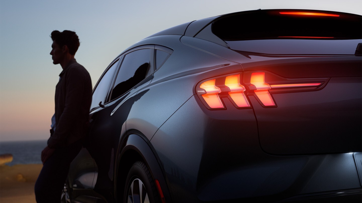 Ford Mustang Mach-E in Schwarz. Dreiviertel-Heckansicht in der Abenddämmerung, ein Mann lehnt an der Fahrertür Ford Mustang Mach-E in Schwarz. Dreiviertel-Heckansicht in der Abenddämmerung, ein Mann lehnt an der Fahrertür