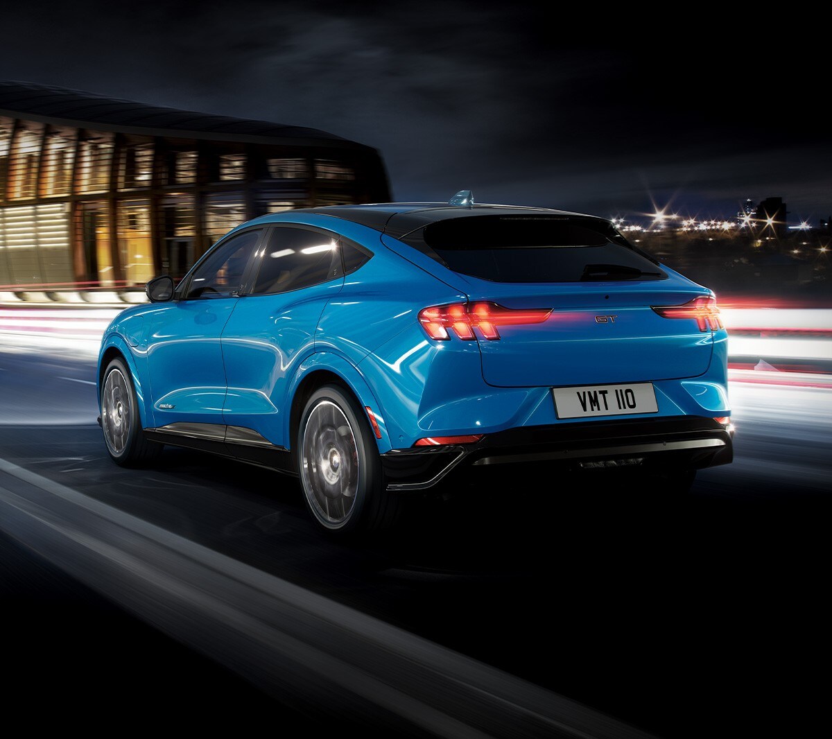 Ford Mustang Mach-E in Blau. Dreiviertel-Heckansicht. Bei Nacht auf einer Straße vor einem modernen gebäude fahrend Ford Mustang Mach-E in Blau. Dreiviertel-Heckansicht. Bei Nacht auf einer Straße vor einem modernen gebäude fahrend