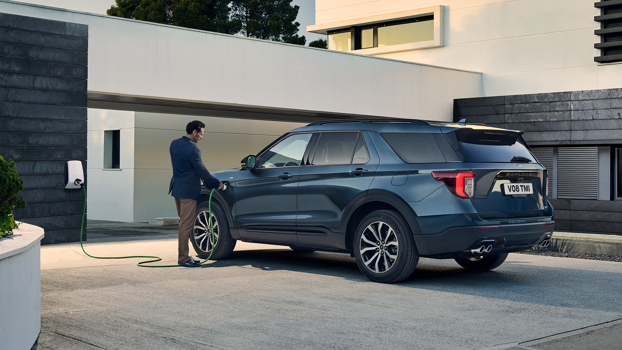 Ford Explorer in Blau in der 7/8 Seitenansicht von hinten parkt vor Familienhaus und wird von Mann an Ladestation angeschlossen 