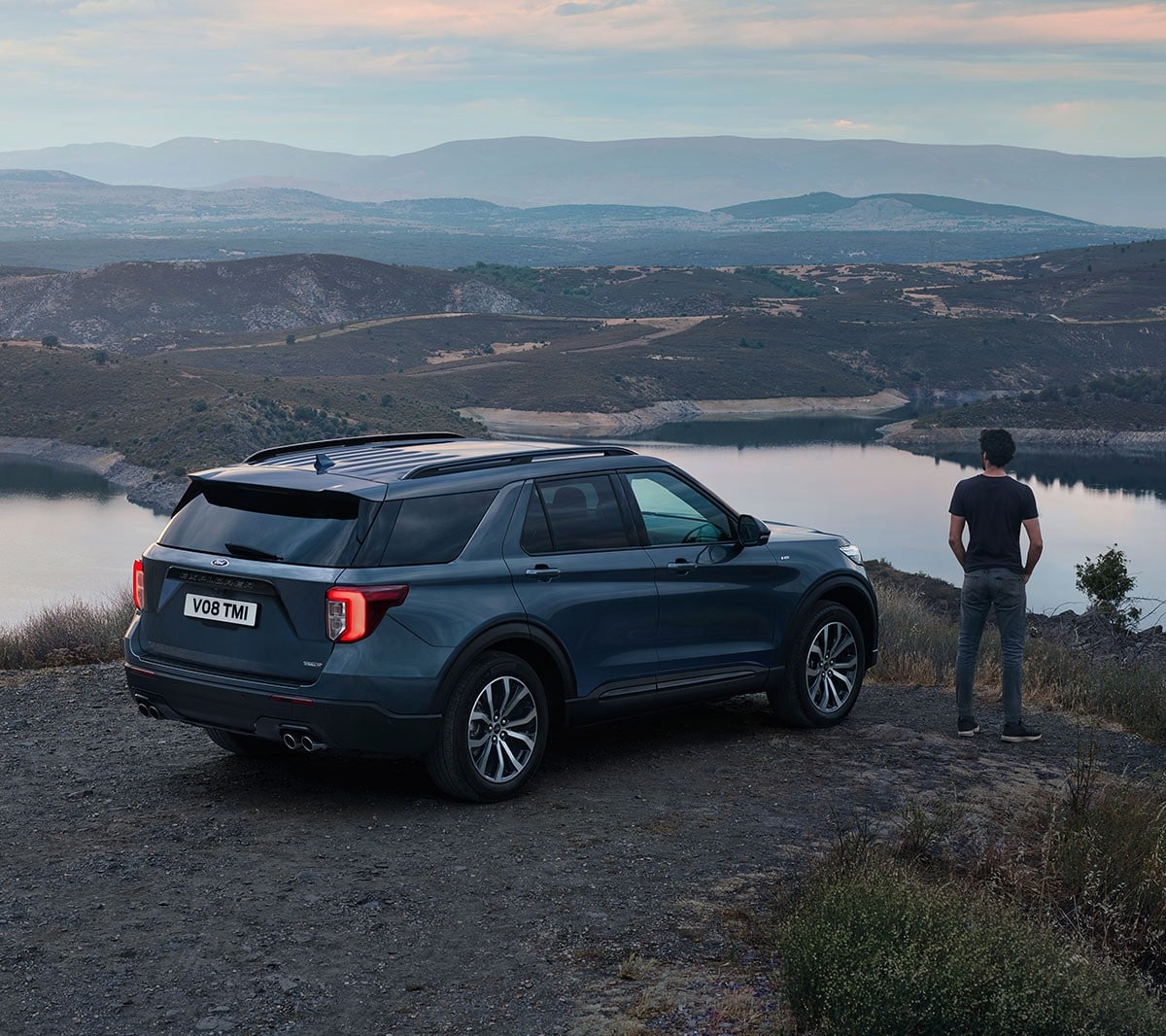 Ford Explorer Plug-in-Hybrid in Blau. Dreiviertel-Heckansicht. In hügeliger Landschaft parkend mit nebenstehender Person die Landschaft betrachtend