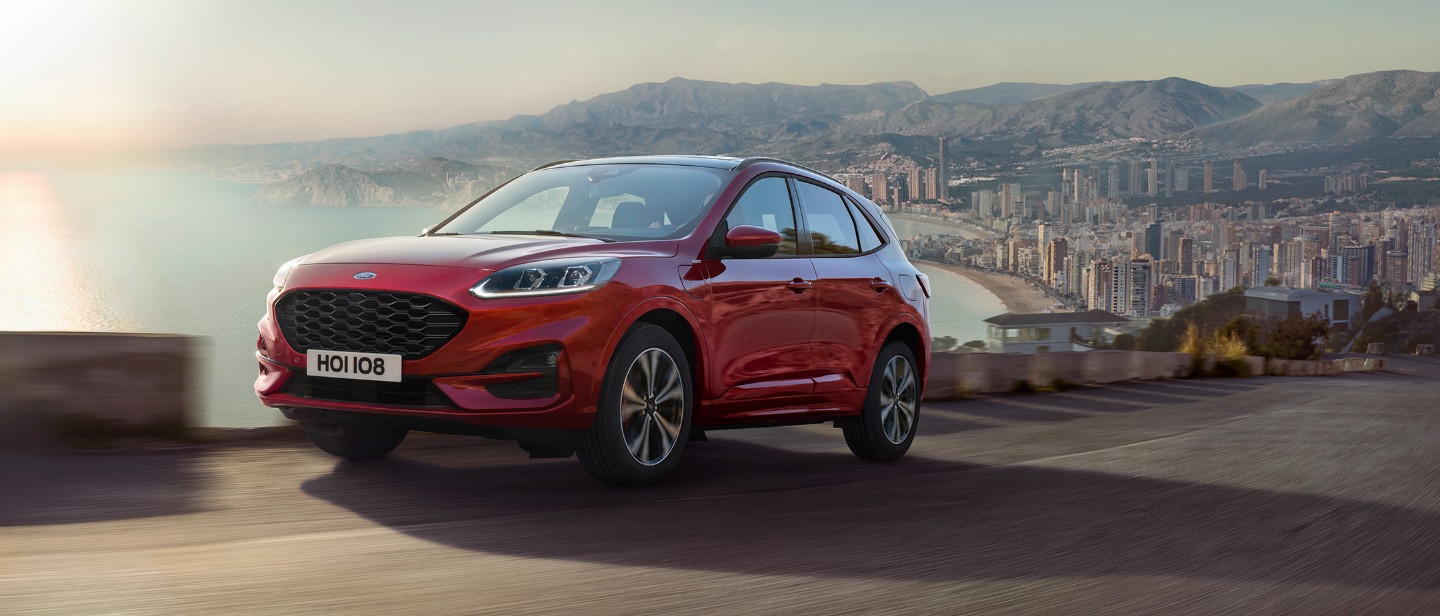 Ford Kuga ¾ Frontansicht in Rot auf einer Straße fahrend vor Küstenpanorama Ford Kuga ¾ Frontansicht in Rot auf einer Straße fahrend vor Küstenpanorama