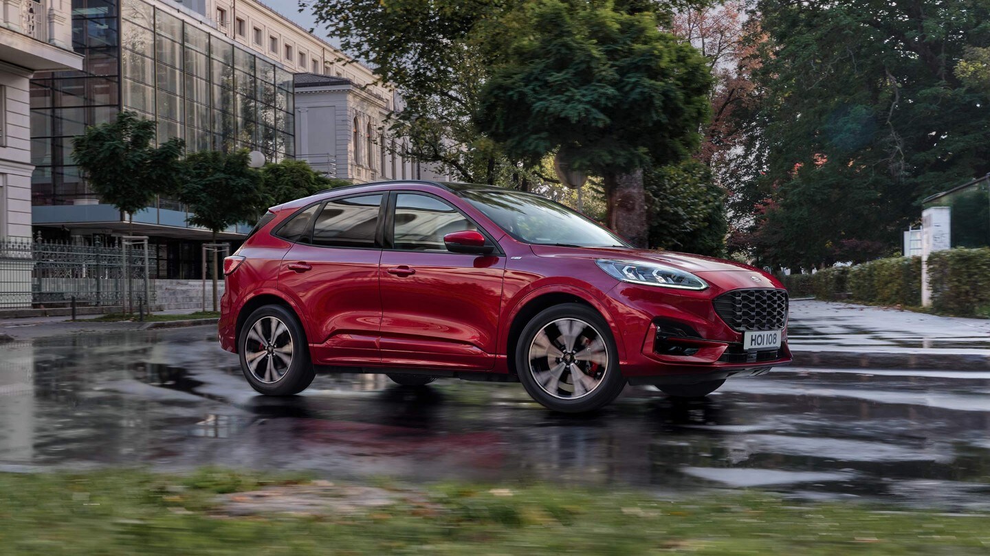 Ford Kuga ¾ Frontansicht in Rot durch eine Stadt fahrend von modernen Gebäuden und Bäumen