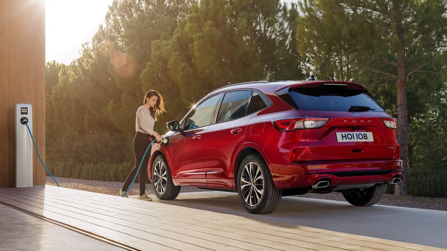 Ford Kuga ¾ Heckansicht in Rot vor Bäumen parkend und eine Frau die das Fahrzeug lädt