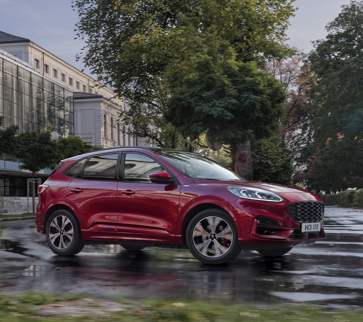 Ford Kuga in Rot. 3/4-Frontansicht, durch eine Stadt vor modernem Gebäude fahrend