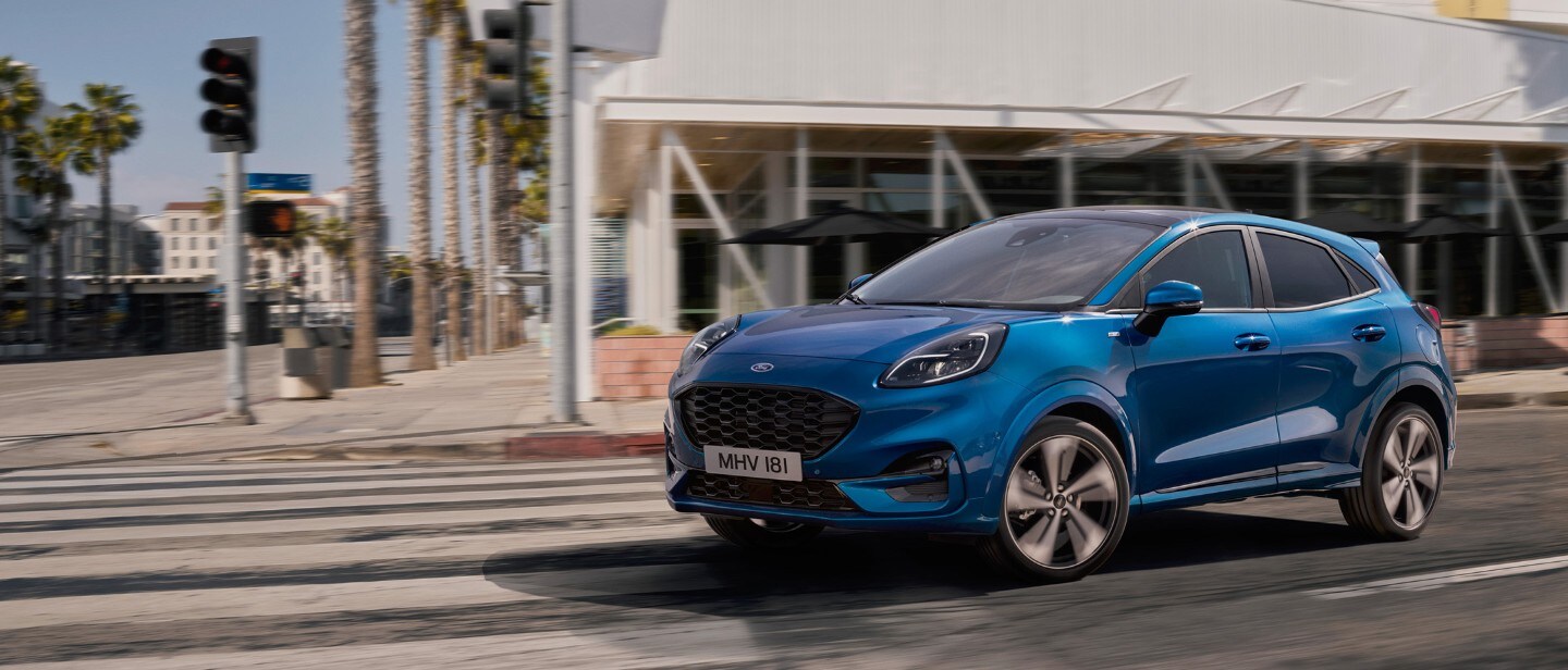 Ford Kuga ¾ Frontansicht in Blau, auf eine Straße fahrend vor einem Gebäude Ford Kuga ¾ Frontansicht in Blau, auf eine Straße fahrend vor einem Gebäude