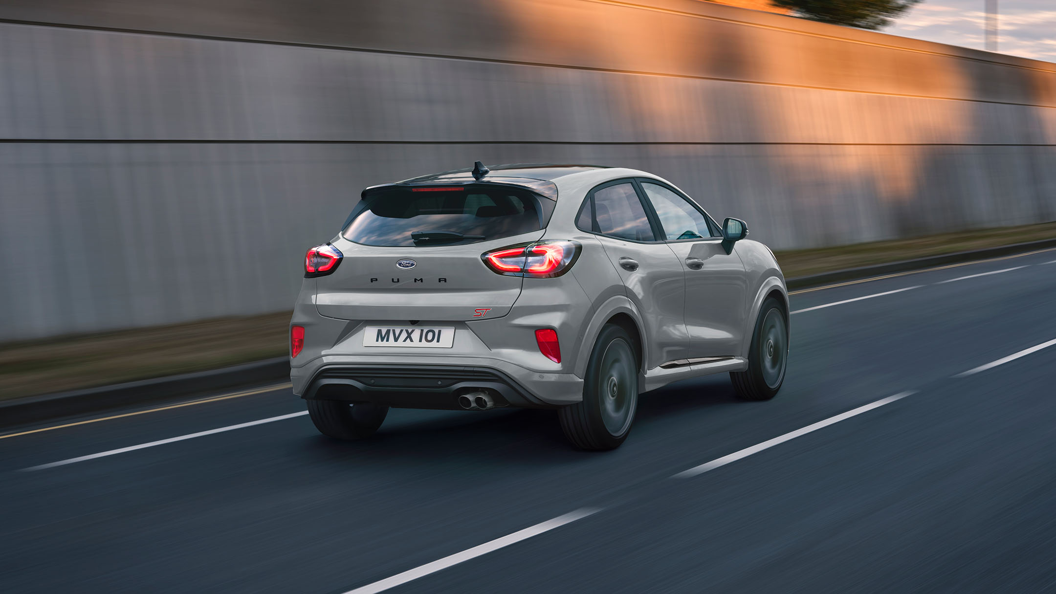Ford Puma ST in Grau. ¾-Frontansicht auf einer Straße vor modernem Gebäude. Eine Frau im Hintergrund