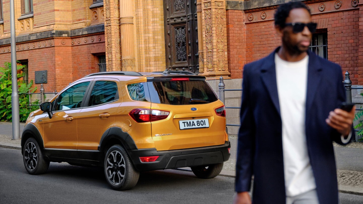 Ford EcoSport in Orange. ¾-Heckansicht, vor einem Gebäude in der Stadt parkend. Mann im Vordergrund schaut auf sein Smartphone