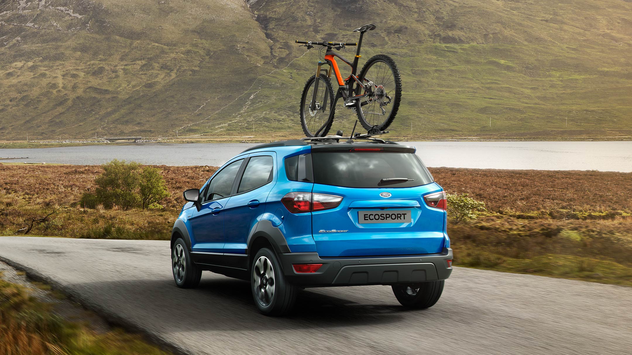 Ford EcoSport in Blau. ¾-Heckansicht. Mit Fahrrad auf dem Dach vor einem See stehend