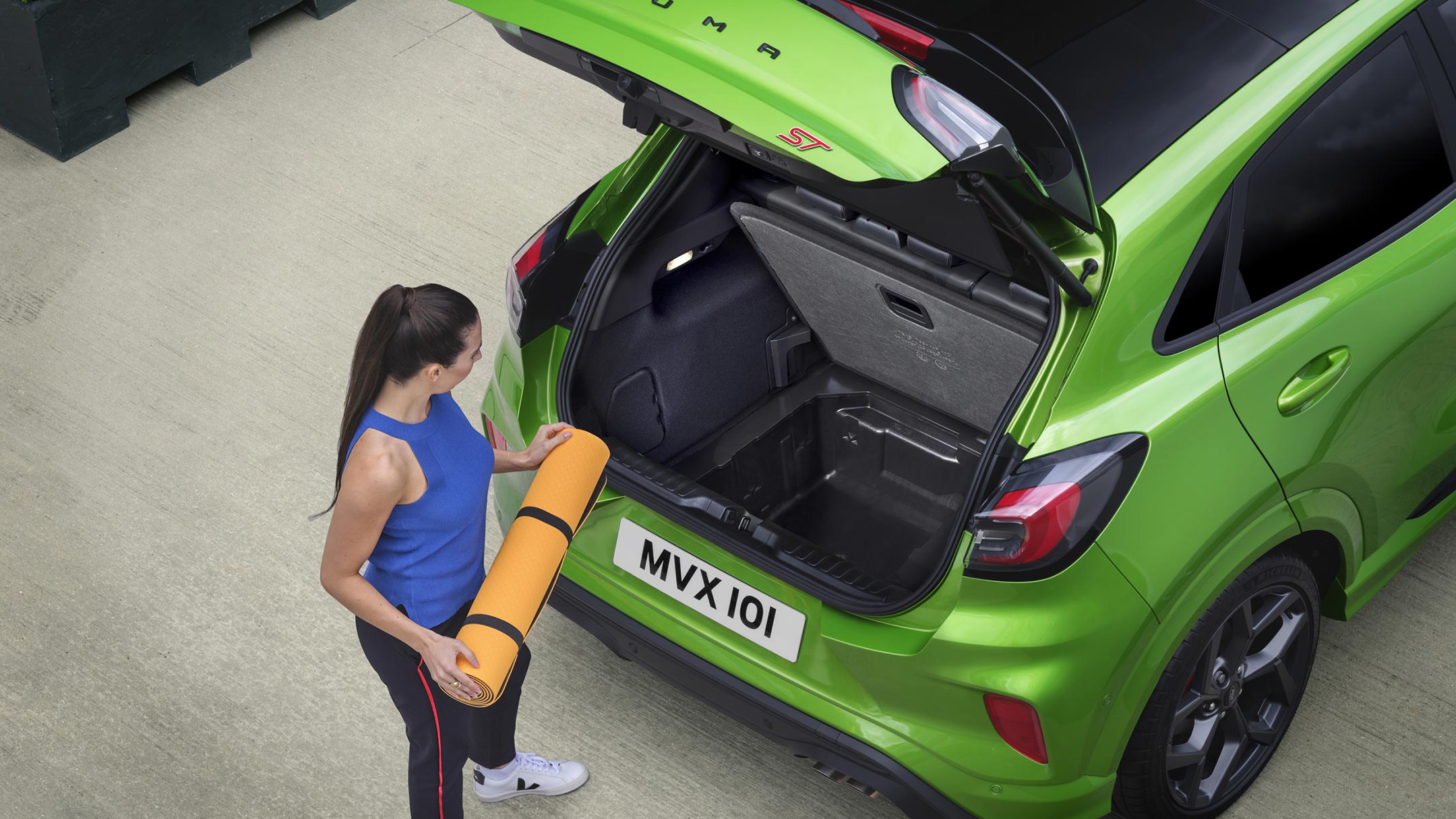 Ford Puma ST in Grün. Dreiviertel-Heckansicht von oben. Frau verstaut eine Matte im offenen Laderaum Ford Puma ST in Grün. Dreiviertel-Heckansicht von oben. Frau verstaut eine Matte im offenen Laderaum