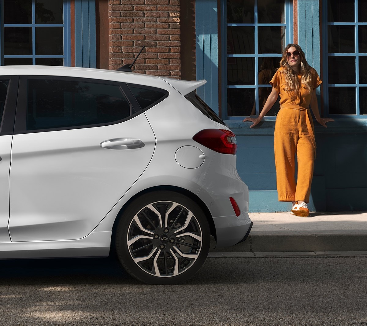 Ford Fiesta in Weiß. Seitenansicht des Hecks, vor einem Haus parkend mit Frau im Hintergrund.
