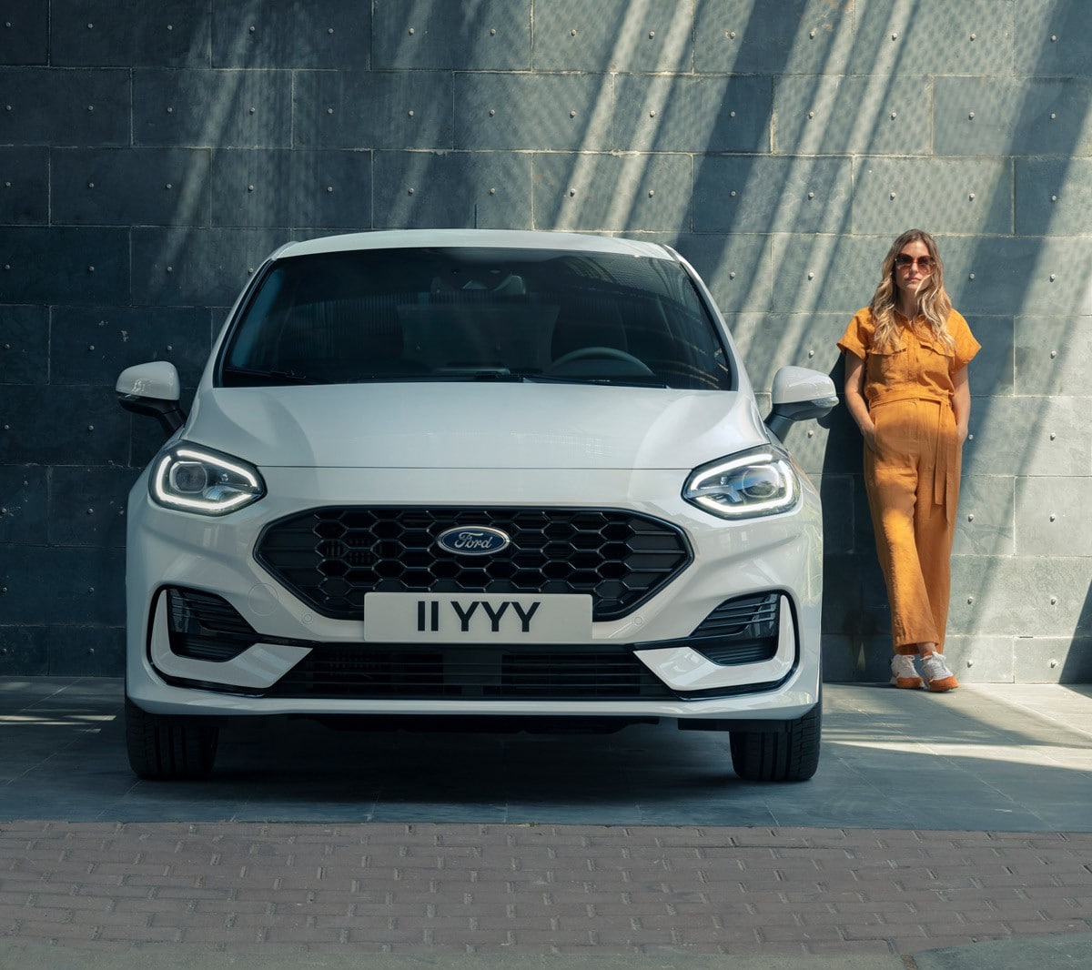 Ford Fiesta in Weiß. Frontansicht vor einem modernen Gebäude parkend mit Frau im Hintergrund.