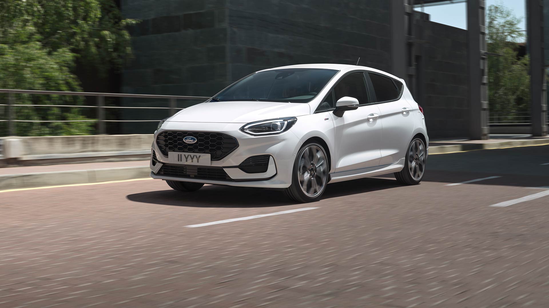 Weißer Ford Fiesta fährt auf einer Stadtstraße, im Hintergrund sind Grünflächen und Gebäude zu sehen.