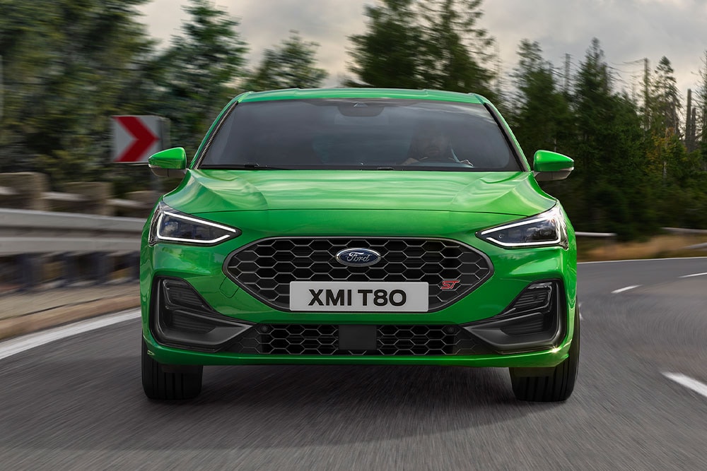 Die Vorderseite eines Ford Focus ST in Mean Green, der um eine Kurve fährt, mit verschwommenen Bäumen im Hintergrund.