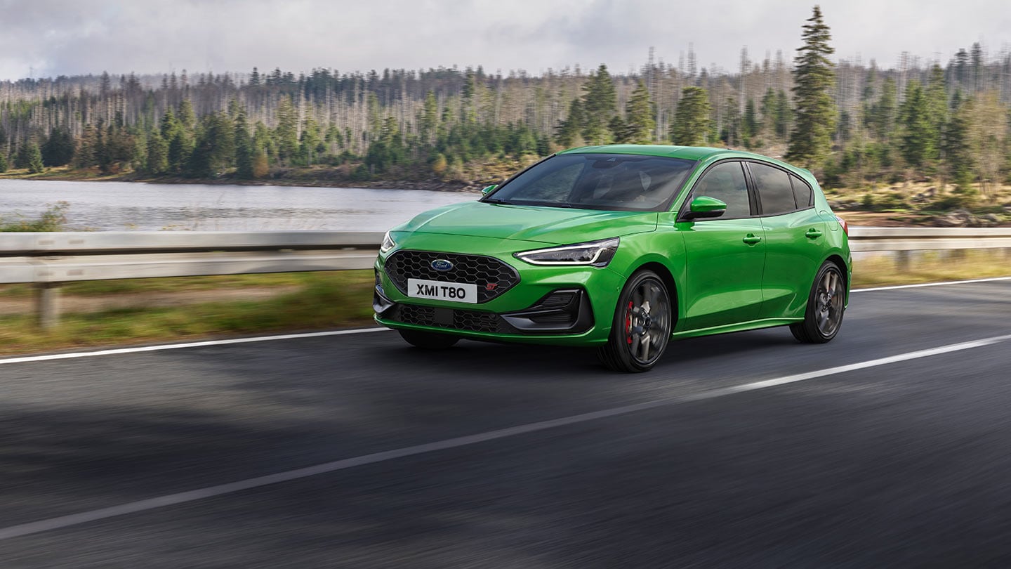 Ein Ford Focus ST in der Farbe "Mean Green" fährt auf einer Straße, mit Bäumen im Hintergrund.