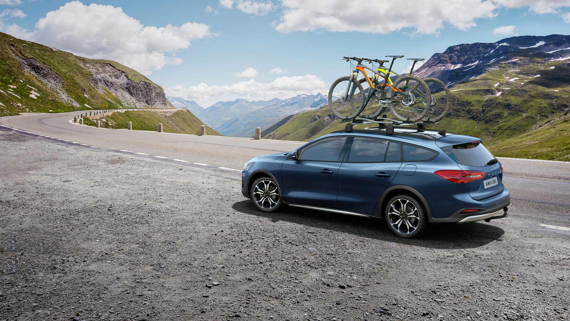 Blauer Ford Focus mit Fahrrädern auf dem Dach fährt entlang einer malerischen Bergstraße.