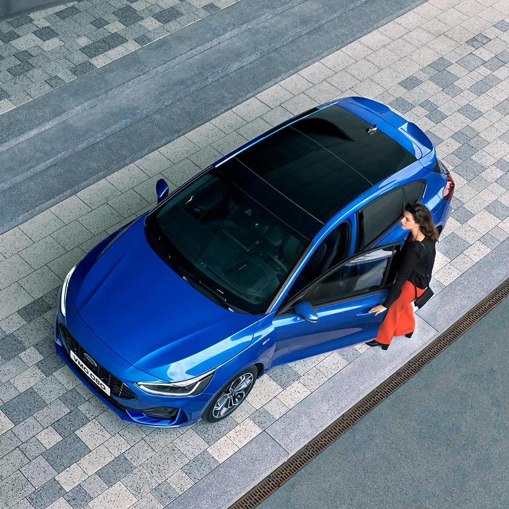 Aus der Vogelperspektive sieht man eine Frau, die in ihren blauen Ford Focus mit Glaspanoramadach einsteigt.