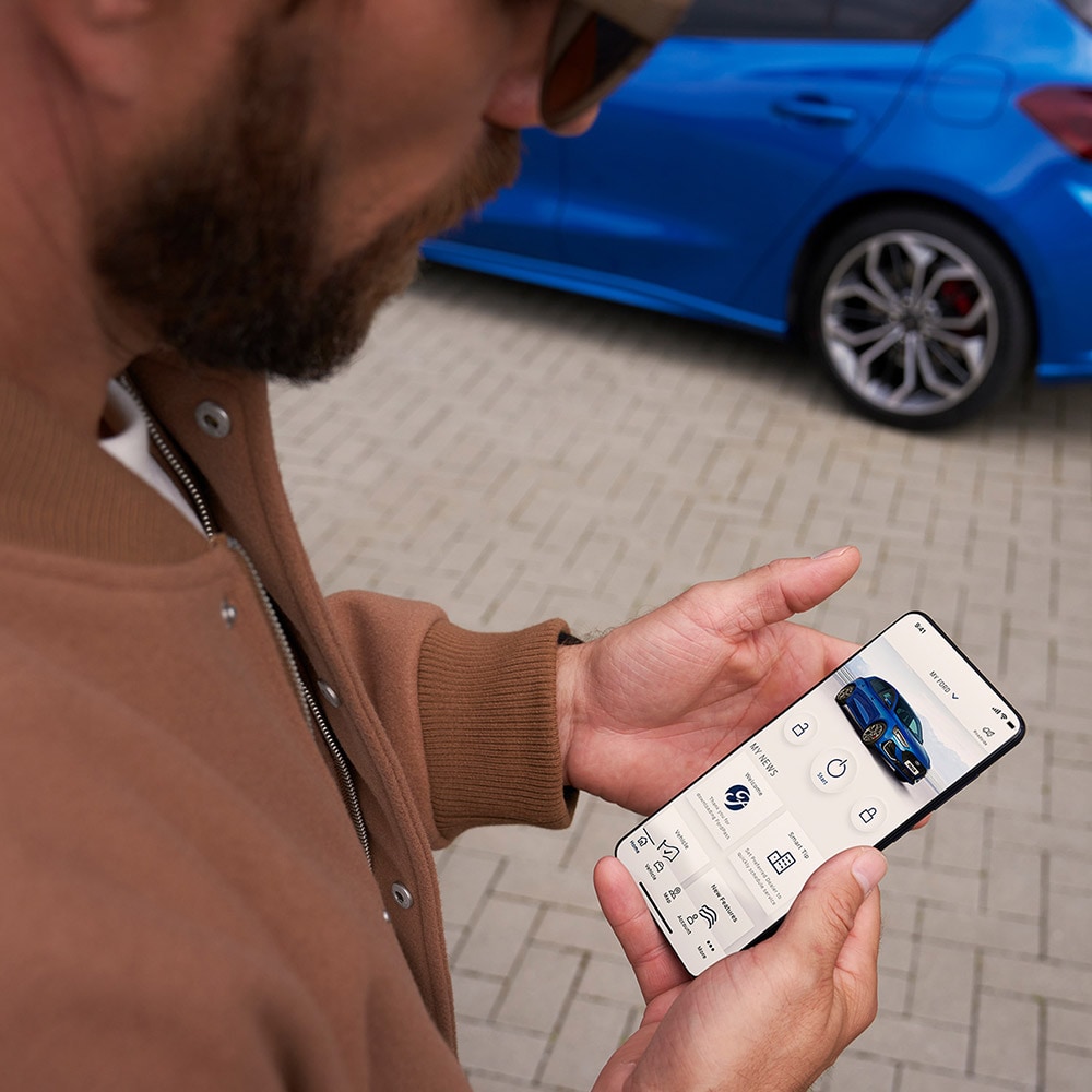 Eine Person hält ein Smartphone, auf dem die FordPass™ App angezeigt wird. Im Hintergrund befindet sich ein blauer Ford Focus.