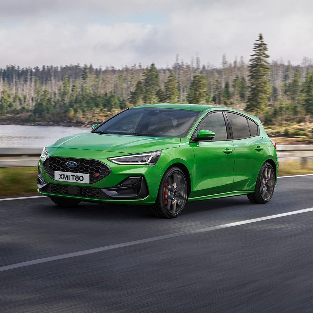 Ein Ford Focus ST in Mean Green fährt auf einer kurvenreichen Straße, umgeben von Bäumen.
