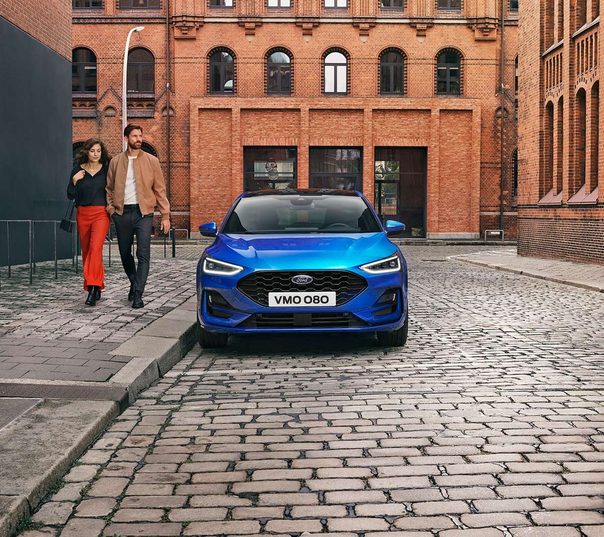 Ford Focus in Blau. Frontansicht, in einer industriellen Gegend parkend. Zwei Personen gehen daneben.