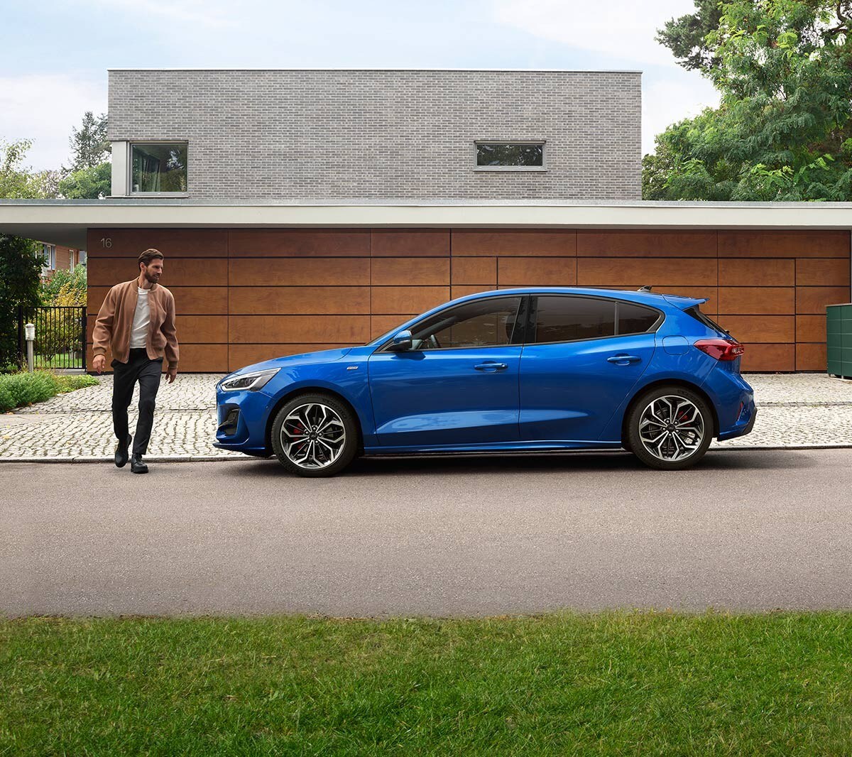 Ford Focus in Blau. Seitenansicht, vor einem Gebäude parkend. Ein Mann läuft daneben.