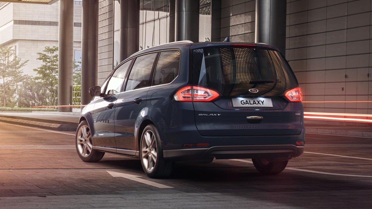 Ford Galaxy Hybrid in Blau. ¾-Heckansicht. Fahrszene vor einem modernen Bürogebäude