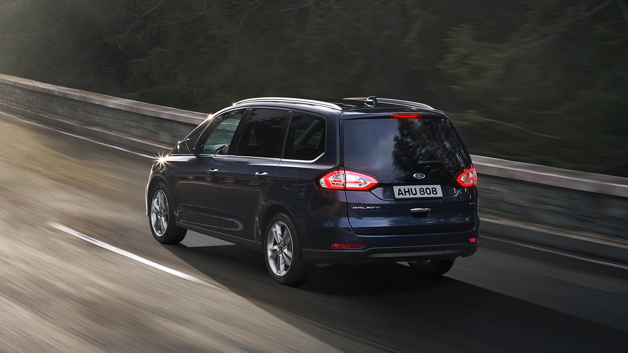 Ford Galaxy Hybrid in Blau. 3/4-Heckansicht. Fahrszene auf einer Landstraße mit Bergen im Hintergrund