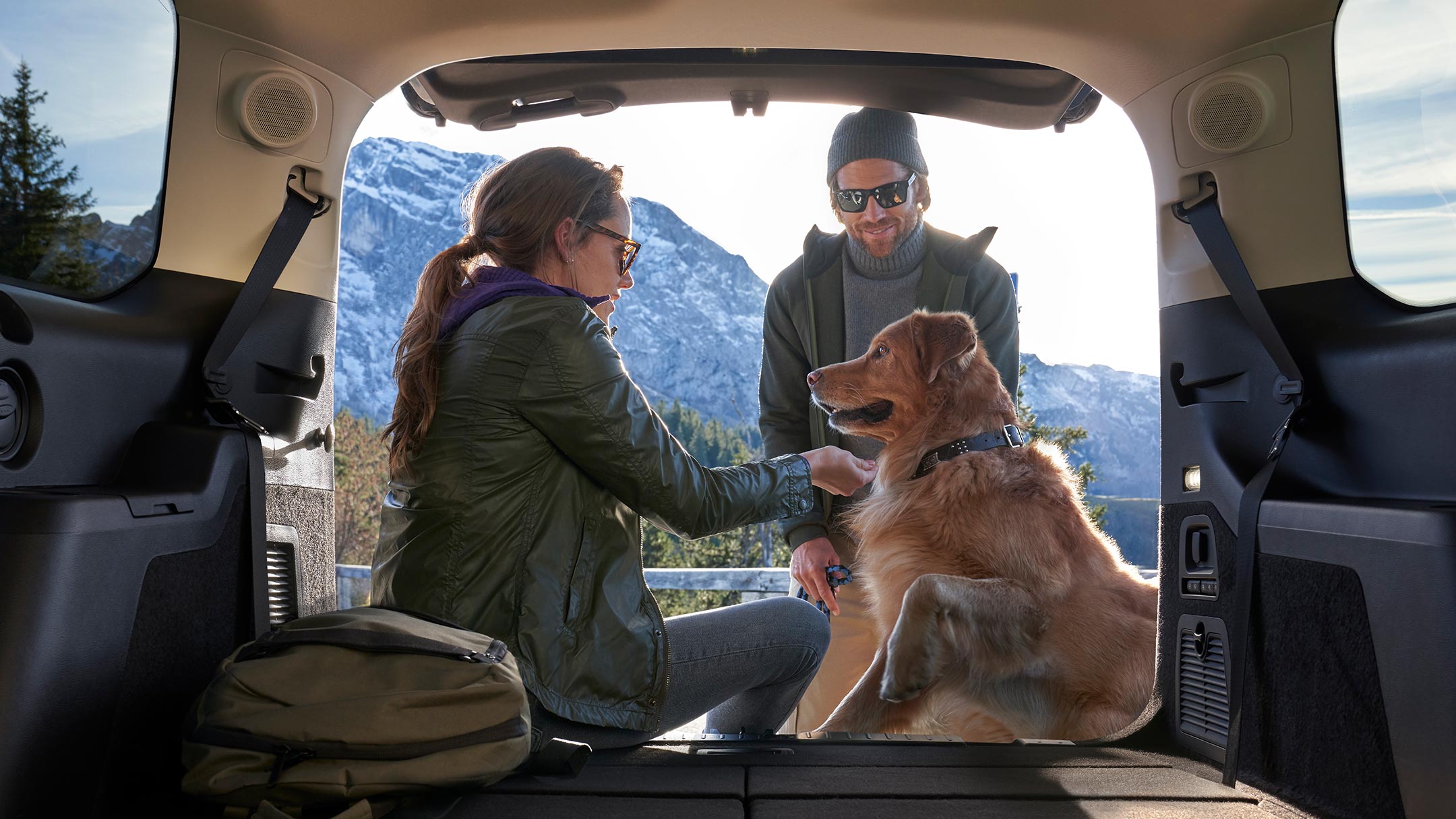 Ford Galaxy Hybrid. Blick von innen durch die geöffnete Heckklappe. Hund und Frau auf der Ladekante sitzend, Mann vor der Heckklappe stehend.