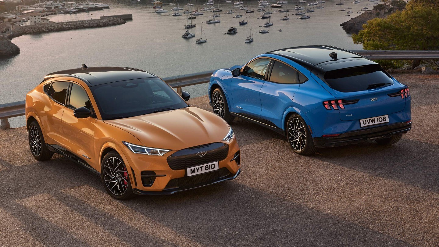 Ford Mustang Mach-E GT. In Orange und Blau. Dreiviertelansicht von oben, vor einem See parkend.