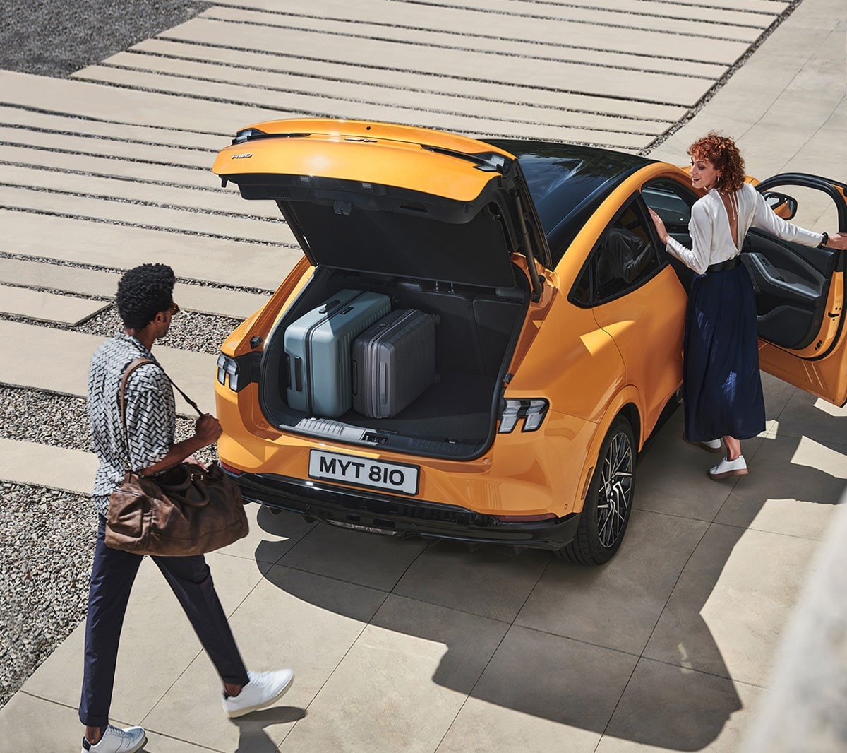 Ford Mustang Mach-E GT in Orange. Dreiviertel-Heckansicht, zwei Personen nutzen die sensorgesteuerte Heckklappe.