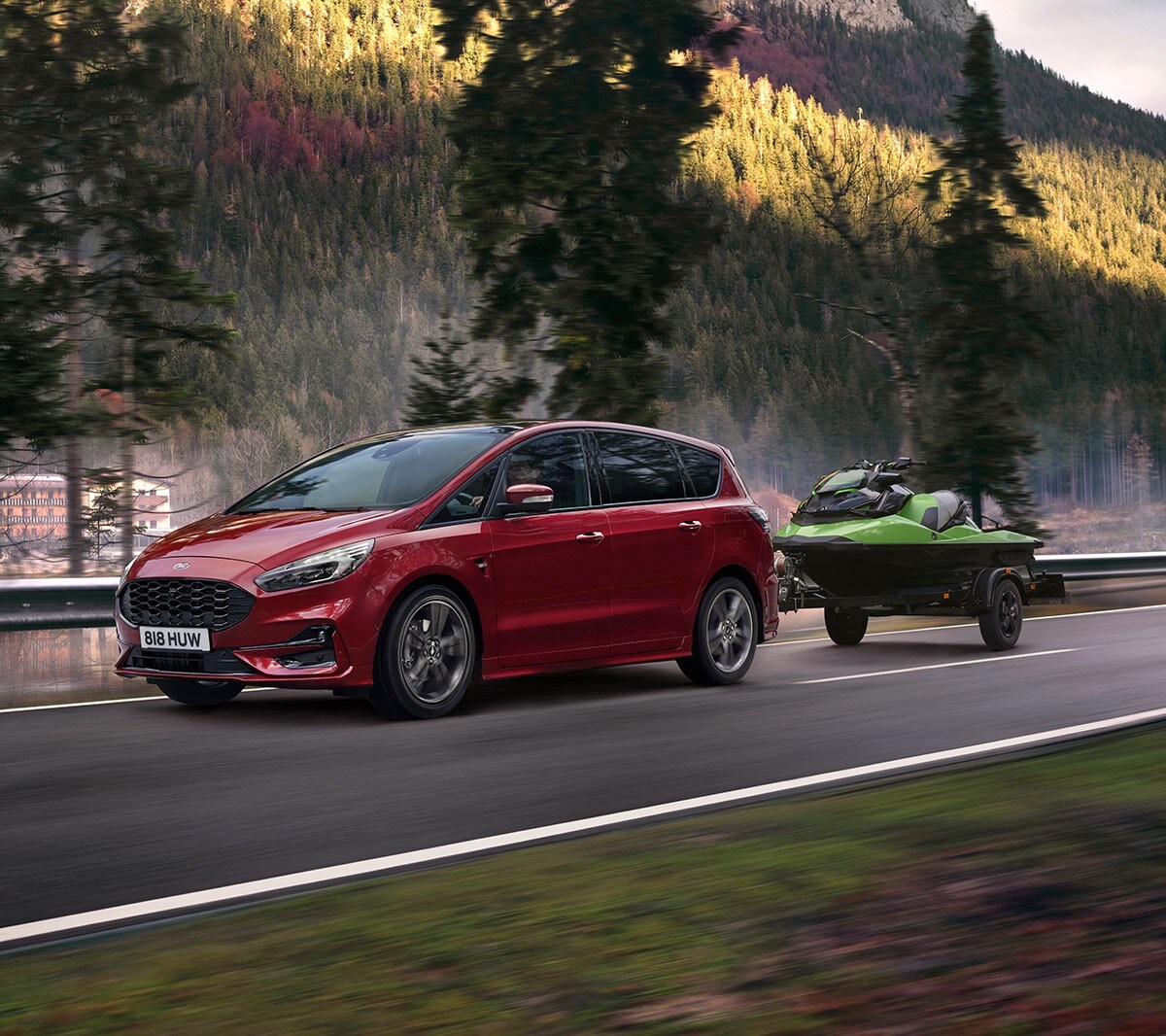 Ford S-MAX Hybrid in Rot. Dreiviertel-Frontansicht, auf einer Straße an einem Fluss mit einem angehängten Jetski fahrend