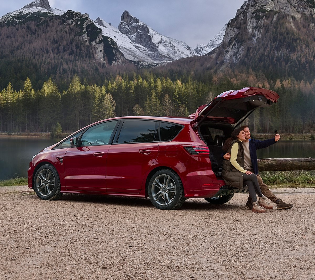 Ford S-MAX Hybrid in Rot. Dreiviertel-Heckansicht, an einem See parkend. Zwei Personen sitzen am Kofferraum