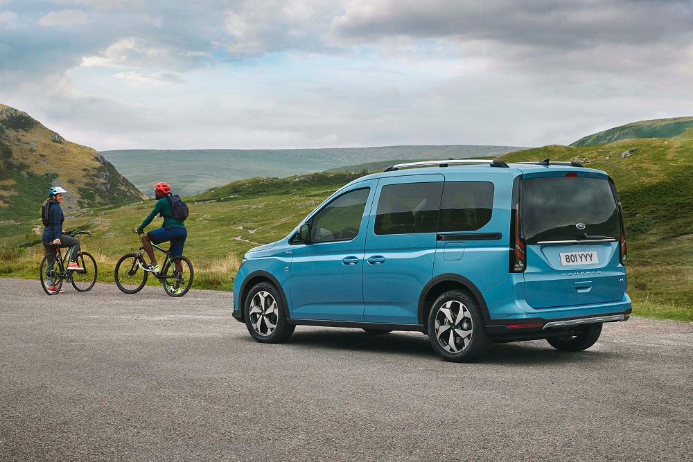 Ein blauer Ford Tourneo Connect parkt auf einer Straße in hügeliger Landschaft mit zwei Radfahrern.