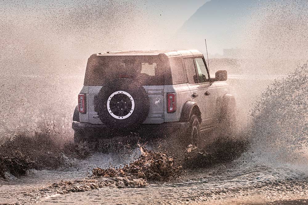Eine Rückansicht eines silbernen Ford Bronco, der durch einen Wasserspray am Strand fährt.