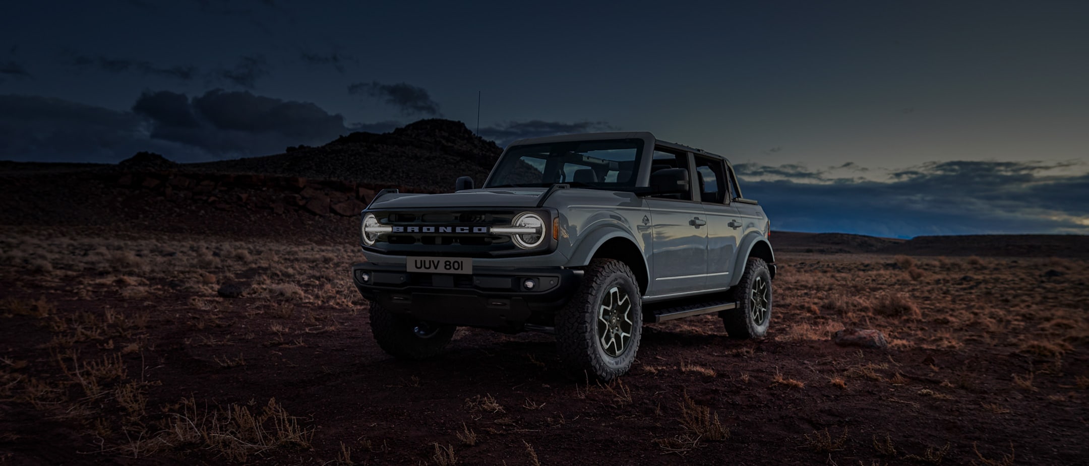 Ford Bronco in Grau. Dreiviertel-Frontansicht, in der Natur im Sonnenuntergang fahrend.