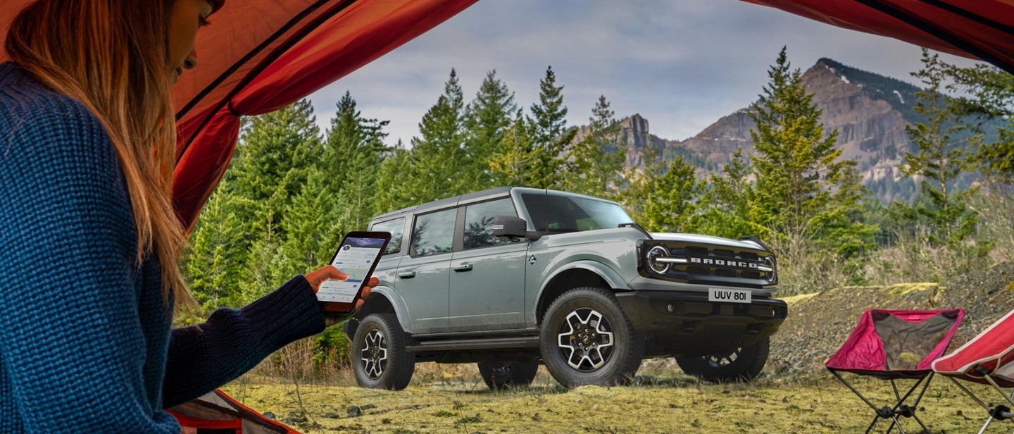 Ford Bronco 3/4 Fontansicht in Grau, vor einem Zelt stehend in dem eine Frau mit Smartphone sitzt