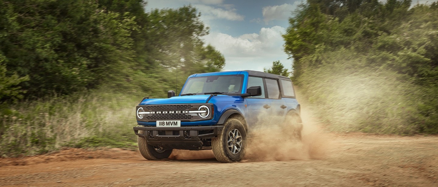 Ford Bronco in Blau,  in 3/4 Frontansicht, auf staubigem Feldweg fahrend