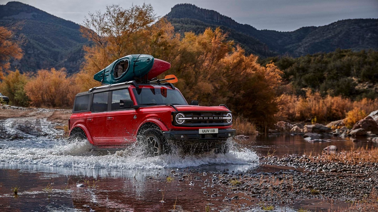 Ford Bronco in Rot, in 3/4-Frontansicht in felsiger Landschaft durch einen Wasserlauf fahrend