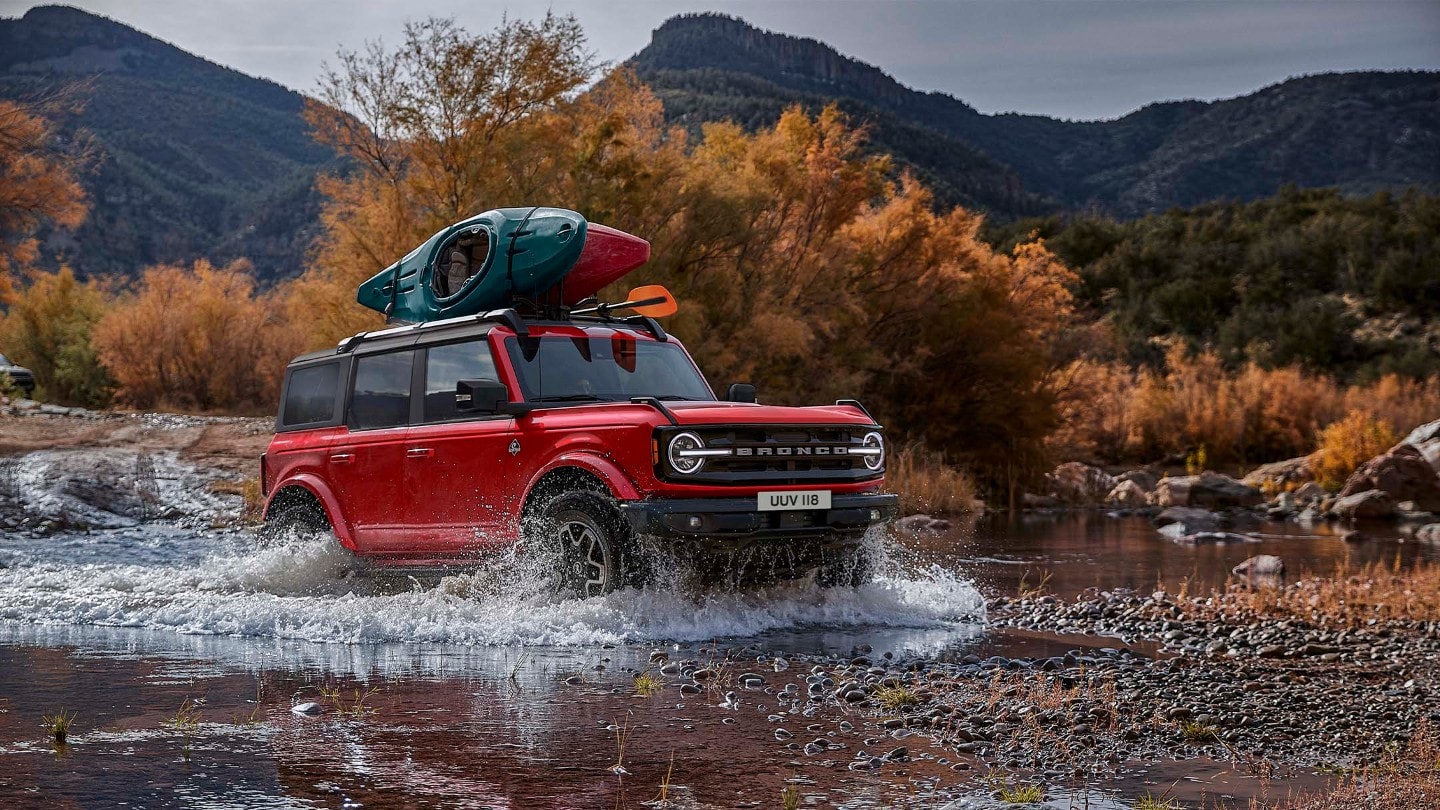 Ford Bronco in Rot. In 3/4-Frontansicht in felsiger Landschaft durch einen Wasserlauf fahrend