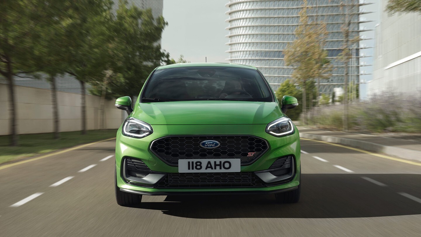 Ford Fiesta ST in Grün. Frontansicht auf Straße vor Bäumen und modernem Gebäude.