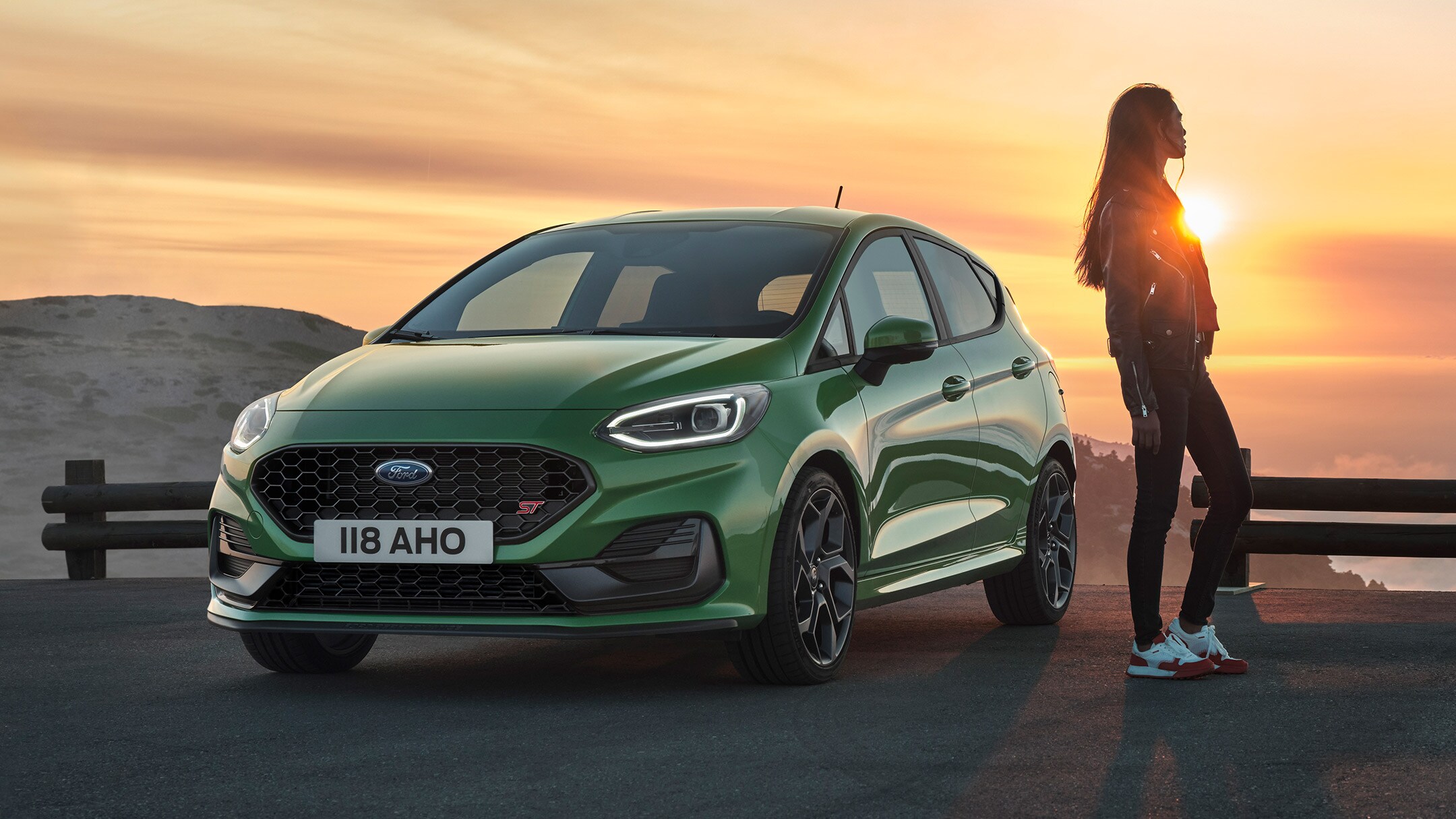 Ford Fiesta ST in Grün. Dreiviertel-Frontansicht, Frau betrachtet den Sonnenuntergang. Ford Fiesta ST in Grün. Dreiviertel-Frontansicht, Frau betrachtet den Sonnenuntergang.