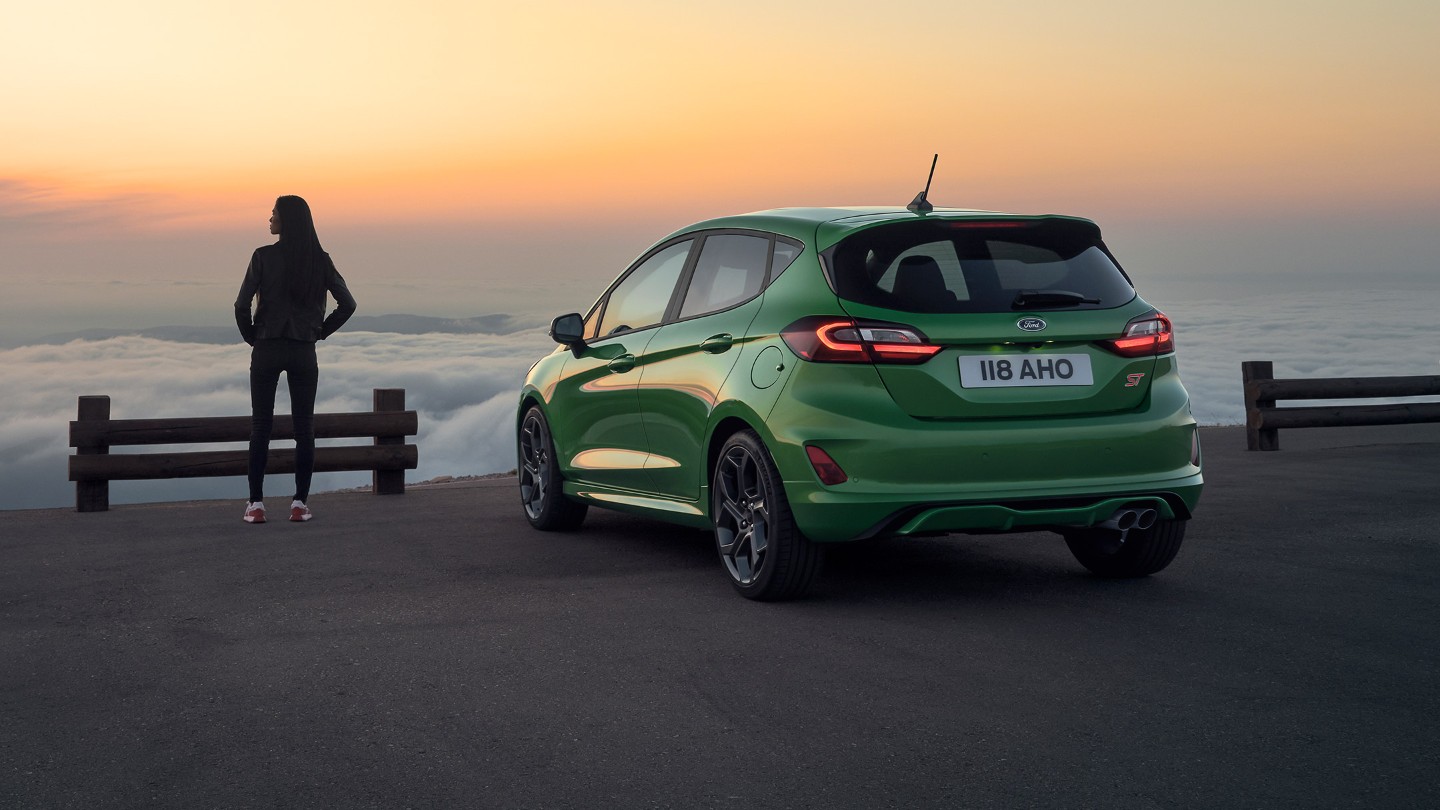 Ford Fiesta ST in Grün. Dreiviertel-Heckansicht, am Meer parkend mit einer Frau im Sonnenuntergang.