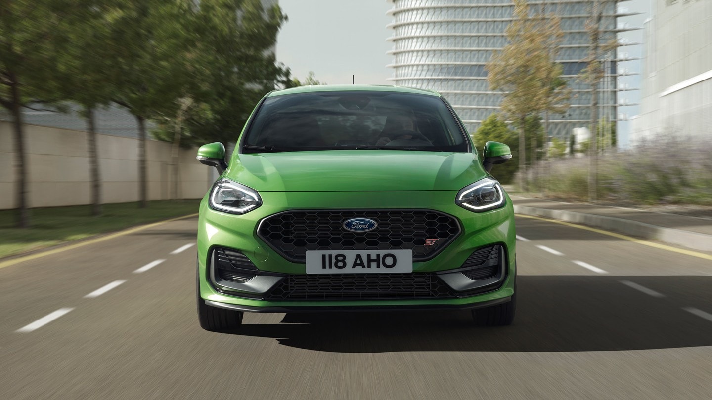 Ford Fiesta ST in Grün. Frontansicht auf Straße vor Bäumen und modernem Gebäude.