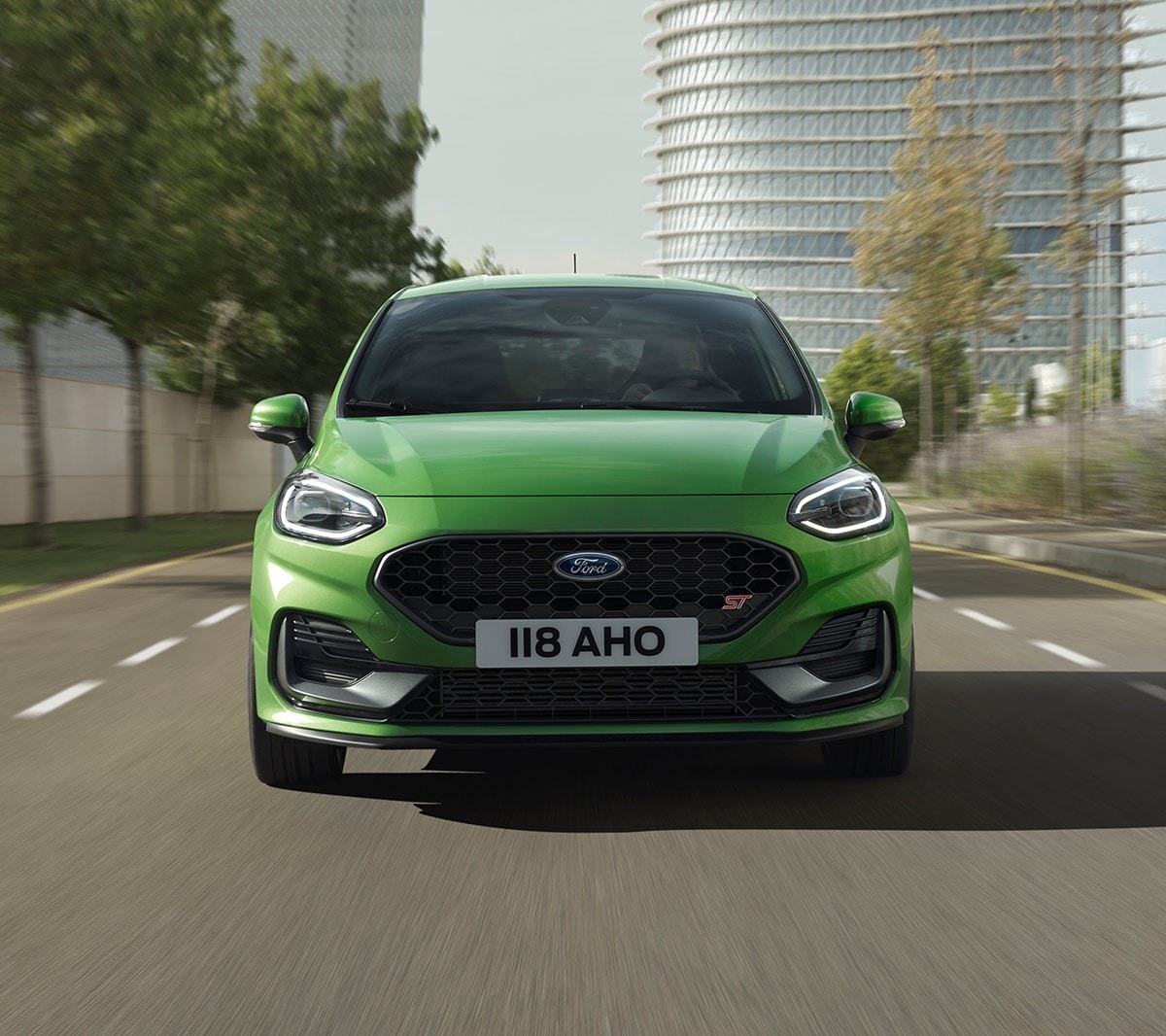 Ford Fiesta ST in Grün. Frontansicht, auf Straße vor Bäumen und modernem Gebäude fahrend.