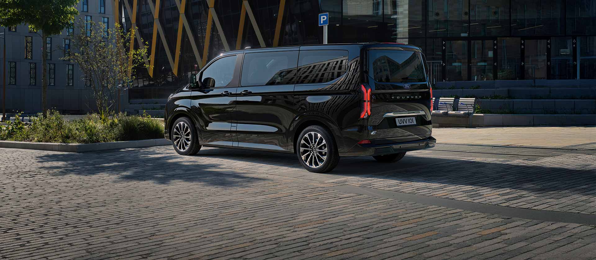 Schwarzer Ford E-Tourneo Custom, Seiten- und Heckansicht vor modernem Gebaeude