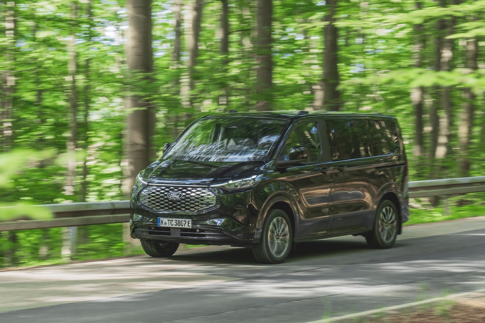 Schwarzer Ford E-Tourneo Custom faehrt auf einer baumbewachsenen Strasse