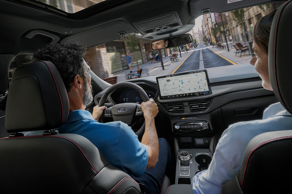 Zwei Personen in einem  Ford Kuga fahren auf einer Stadtstraße und folgen den Anweisungen auf dem 12,3-Zoll-Digitaldisplay.