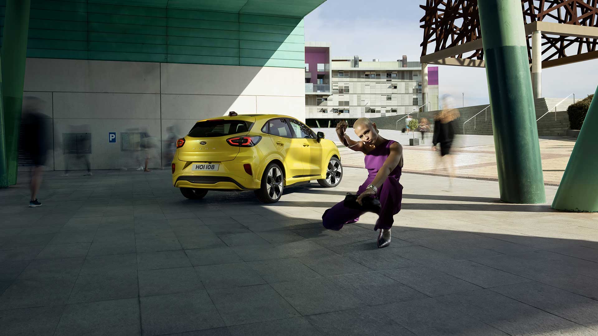 Heckansicht eines gelben elektrischen Ford Puma Gen-E®, der unter einer Brücke parkt, neben einer violett gekleideten Person.