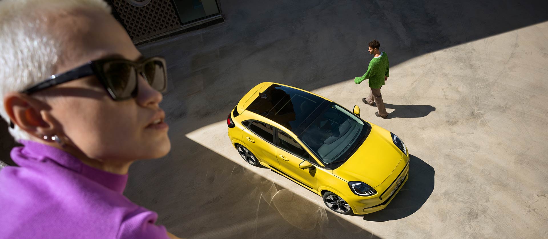 Gelber elektrischer Ford Puma Gen-E® aus der Vogelperspektive. Im Vordergrund ist eine violett gekleidete Person zu sehen. 