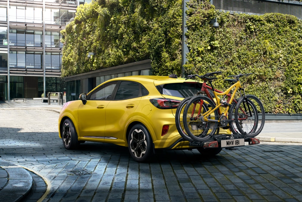 Gelber Ford Puma Gen-E mit zwei Mountainbikes auf einem Fahrradträger, geparkt in einer urbanen Umgebung.